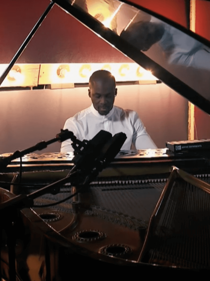 Pianist playing a grand piano in a studio. Black man in white shirt, microphones nearby. Warm lighting.