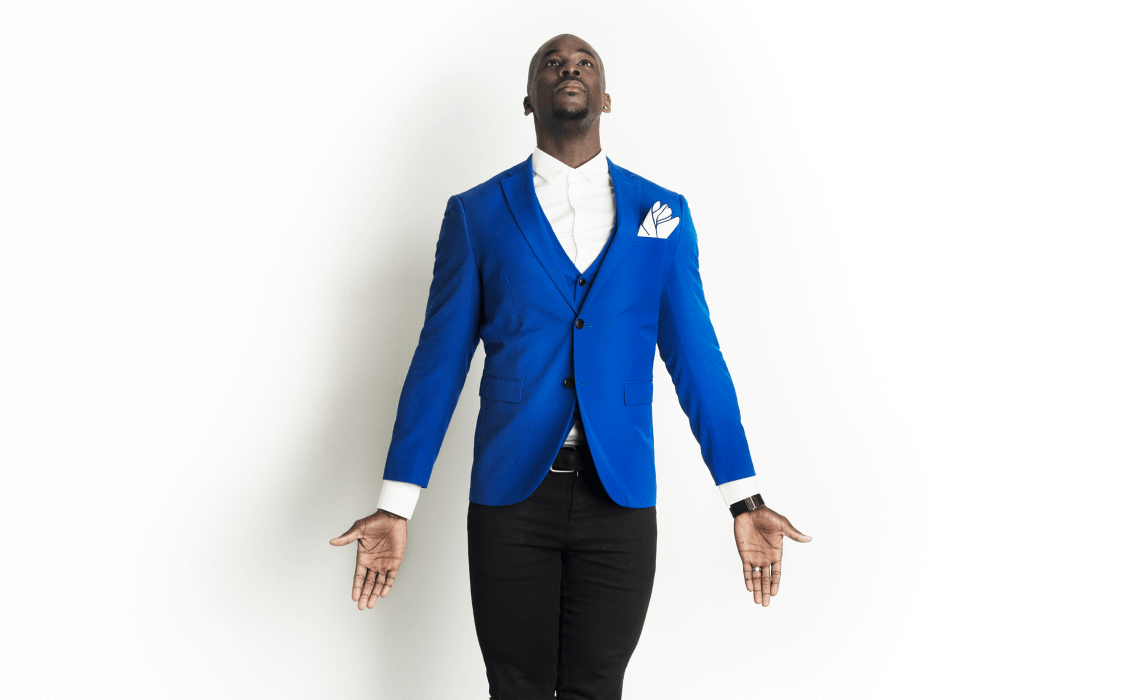 Man in a blue suit looking upward with arms outstretched against a white backdrop.