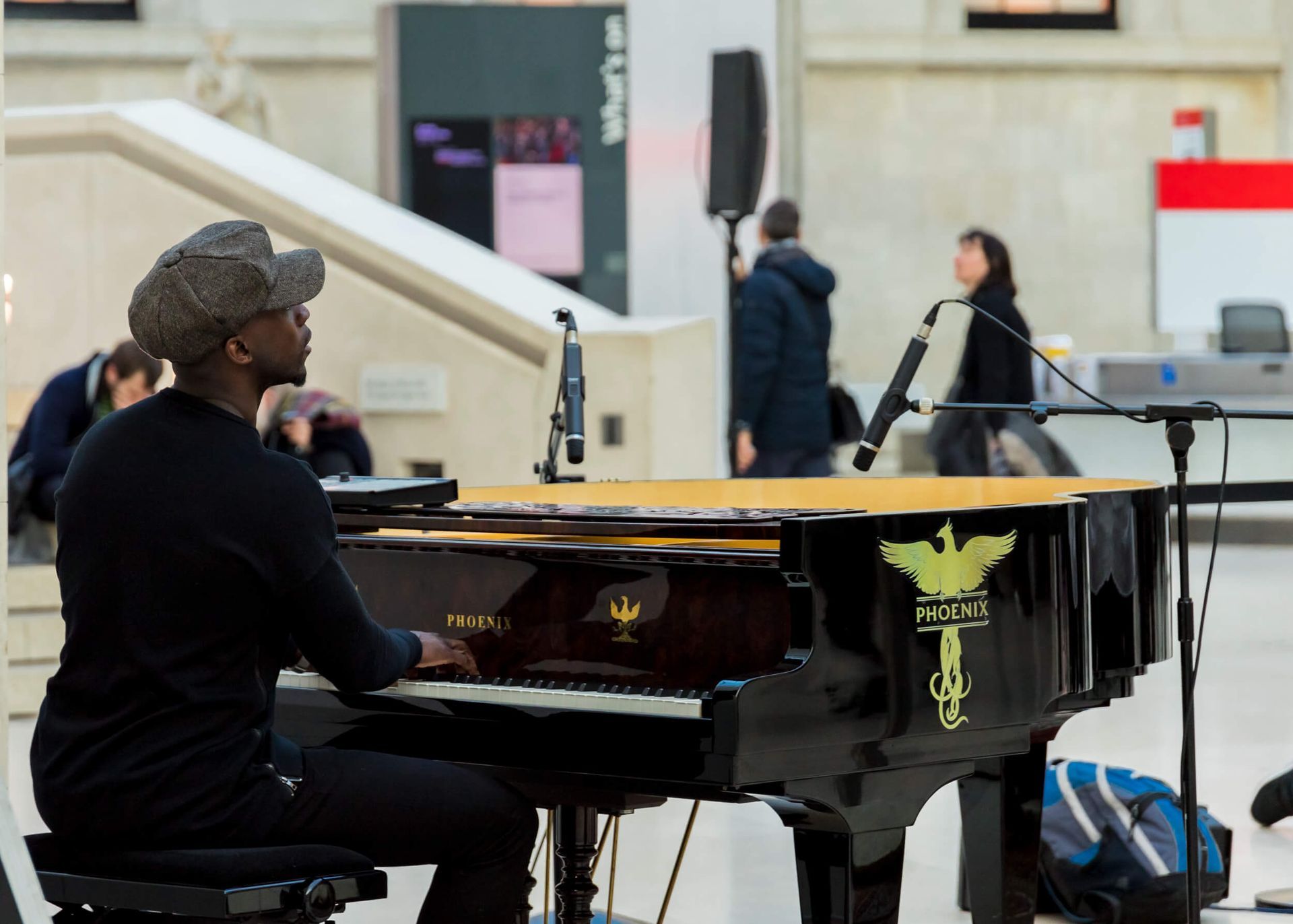 A person playing a black grand piano indoors. They wear a hat and dark clothing. Microphones are nearby.