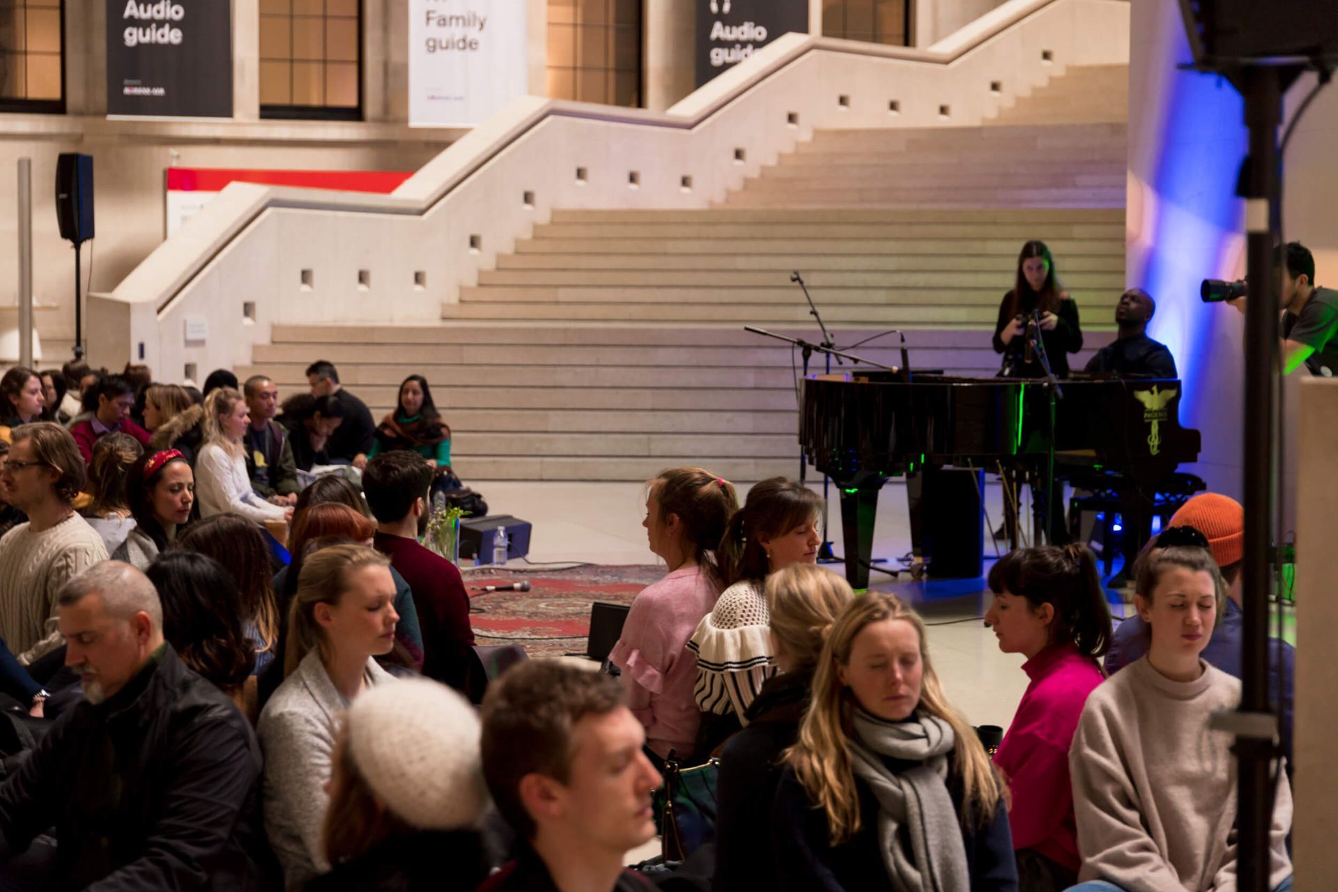 Audience seated, watching a performance on a stage with a piano and musicians in a museum.