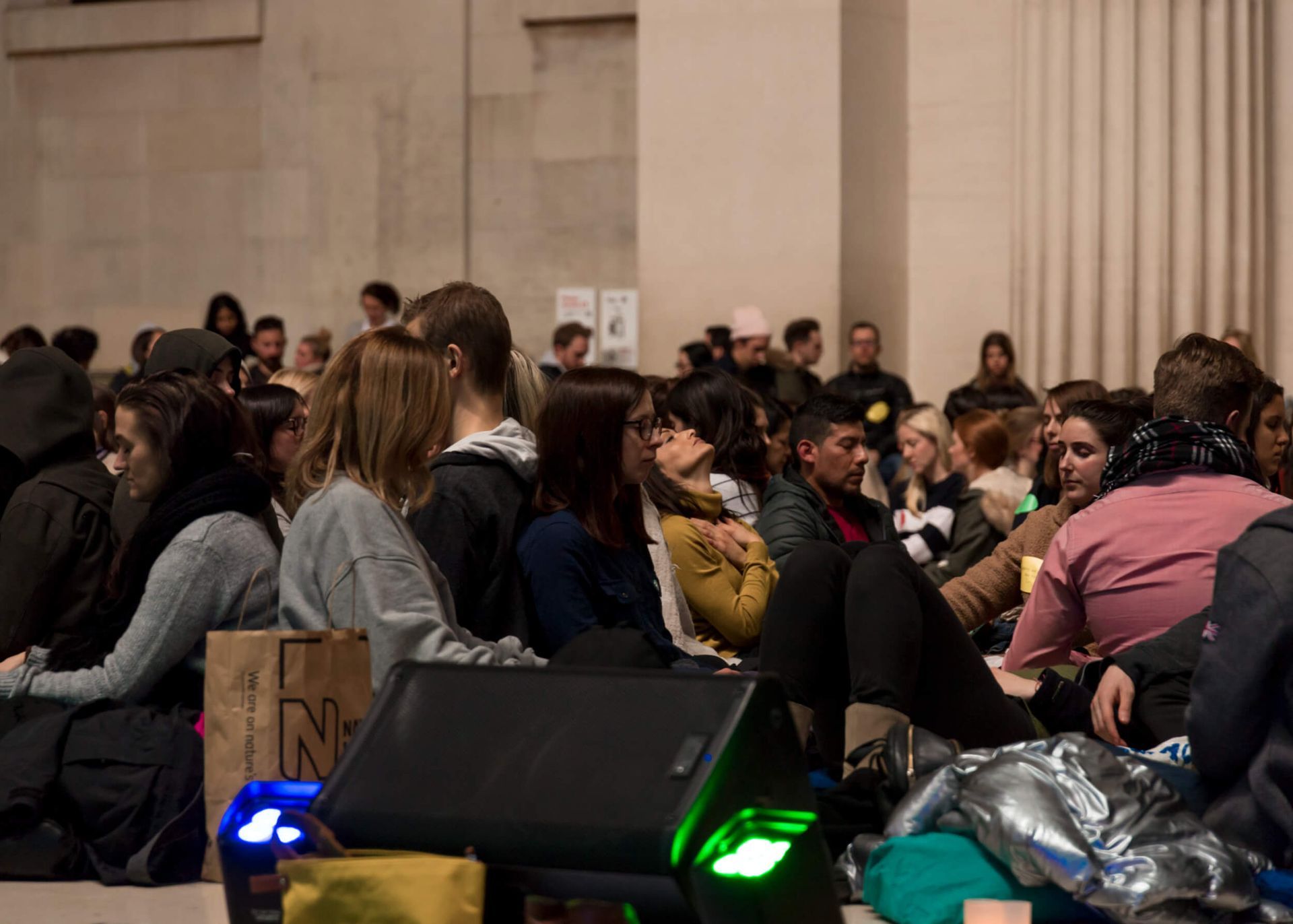 People seated on the floor in a large hall, some talking, others looking forward. Dim lighting.