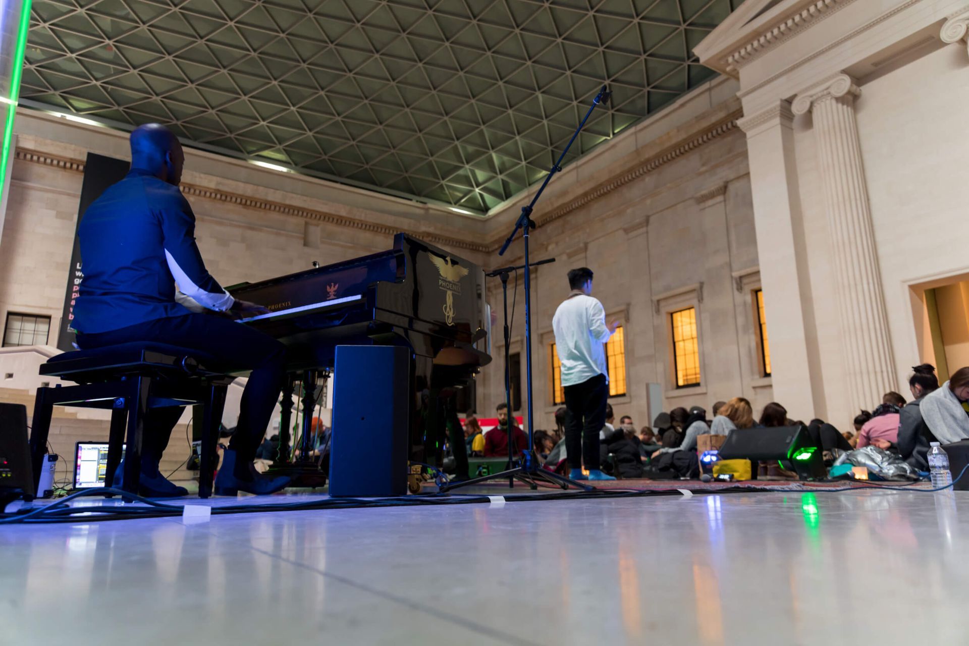 A piano player in blue hoodie and singer performing on stage in a large hall with audience.