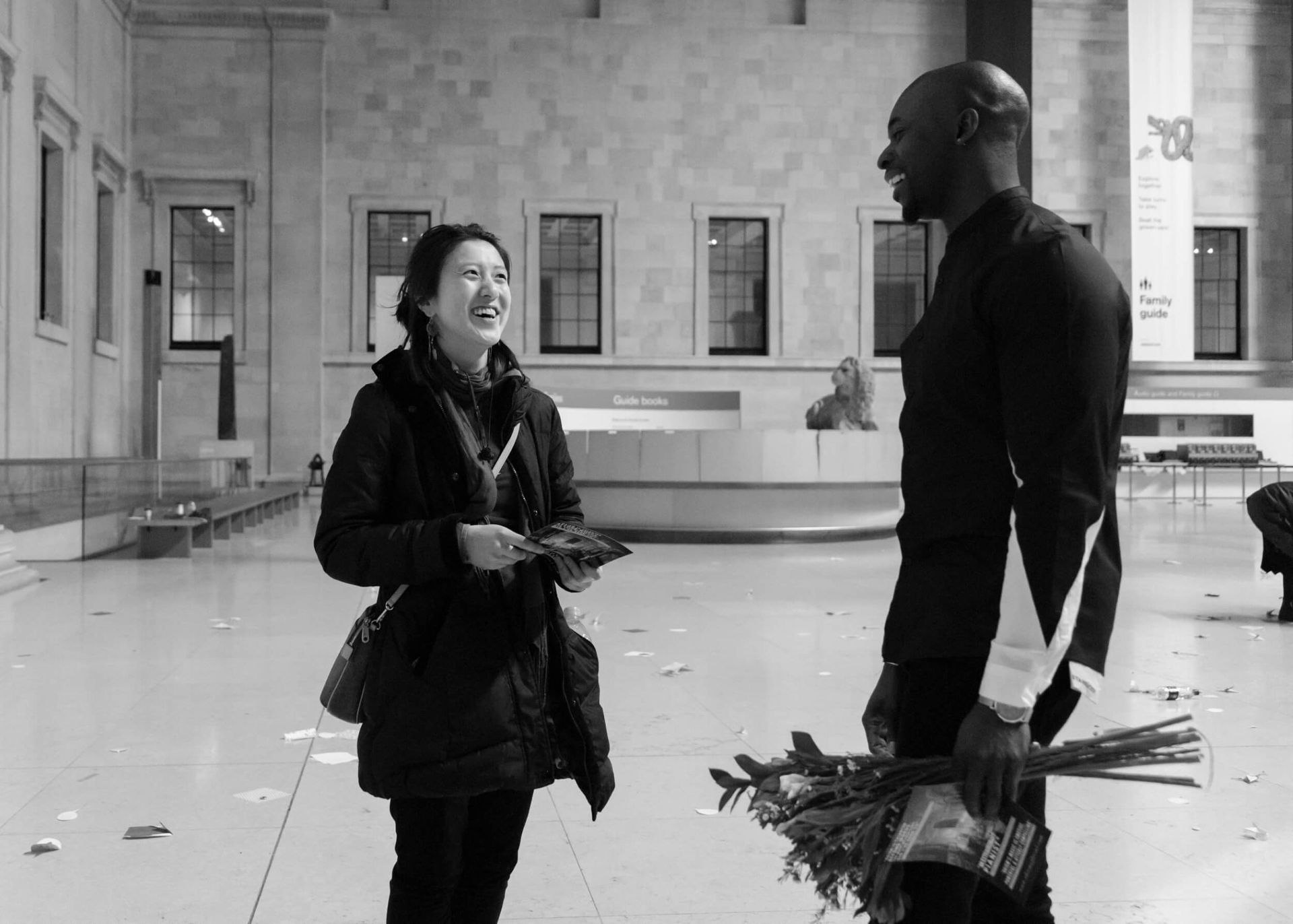 Woman and man laughing, holding flowers, in a large interior space with windows and a reception desk.