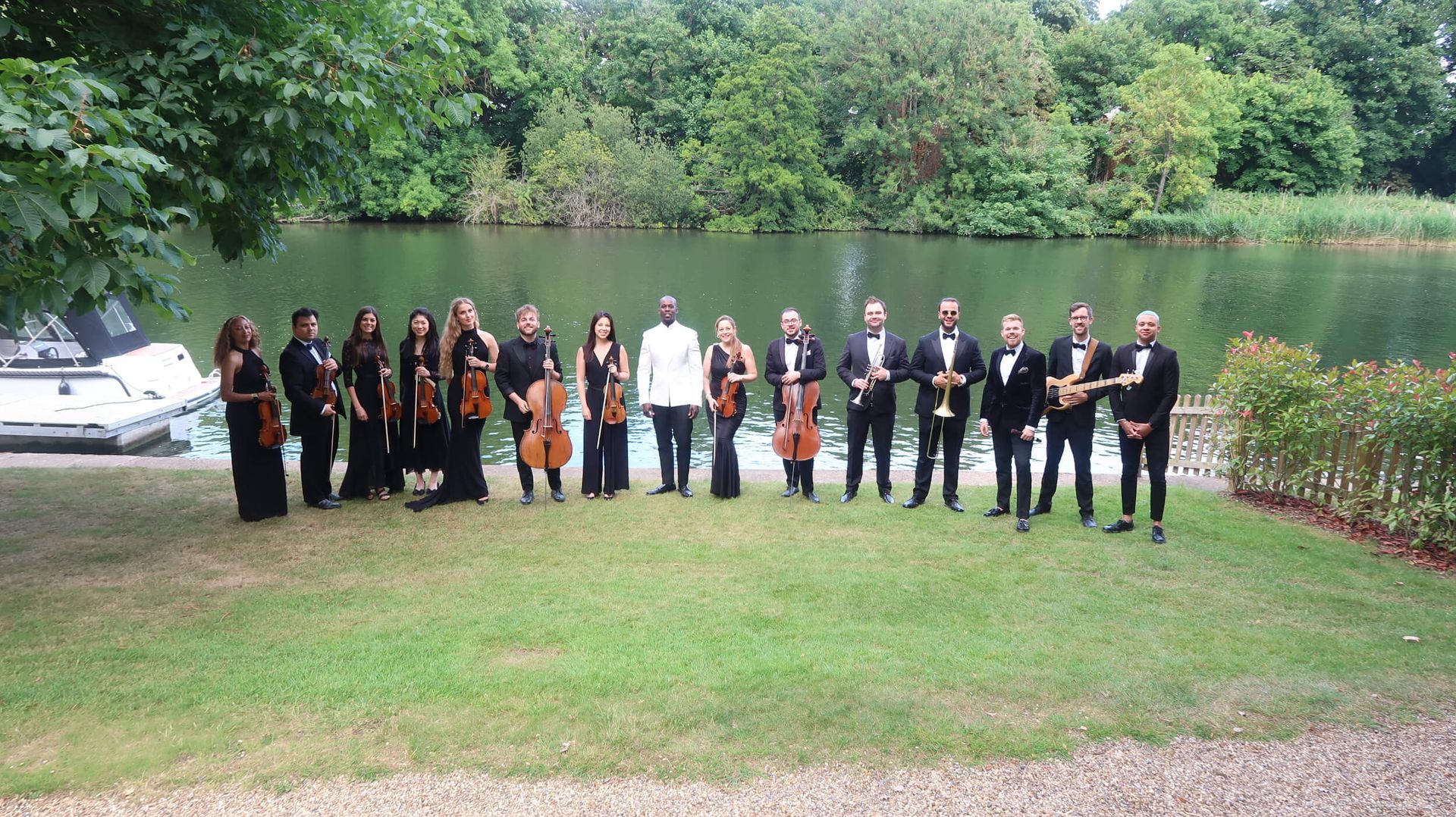 Orchestra standing on grass by a lake, performing. Musicians in formal wear, instruments visible.
