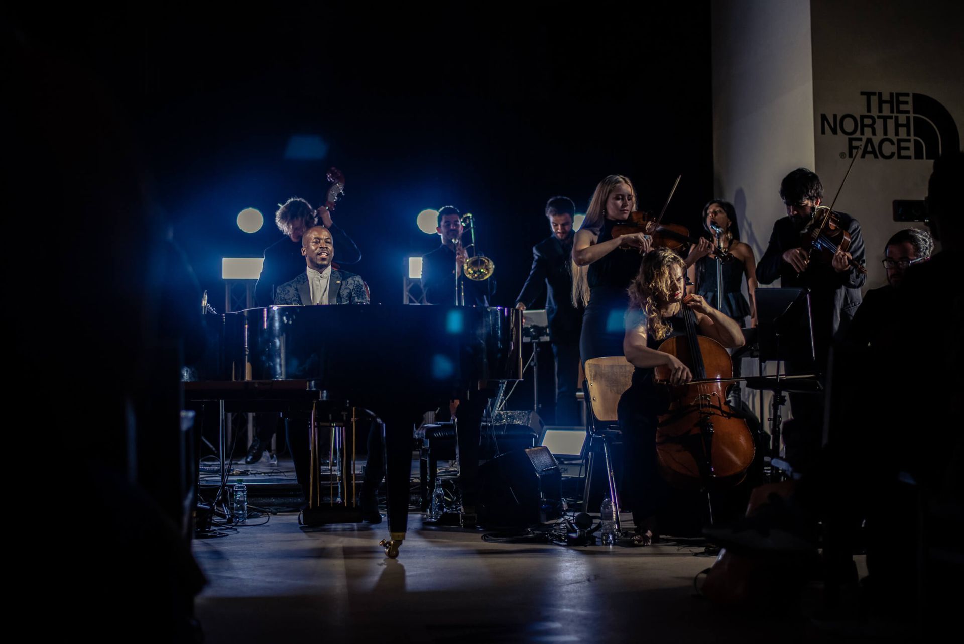 Musicians performing on stage; piano player, string instruments, and brass. Dim lighting, a
