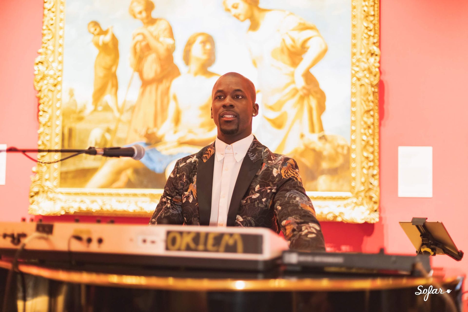 Man playing keyboard at an event, wearing a patterned blazer, in front of a painting.