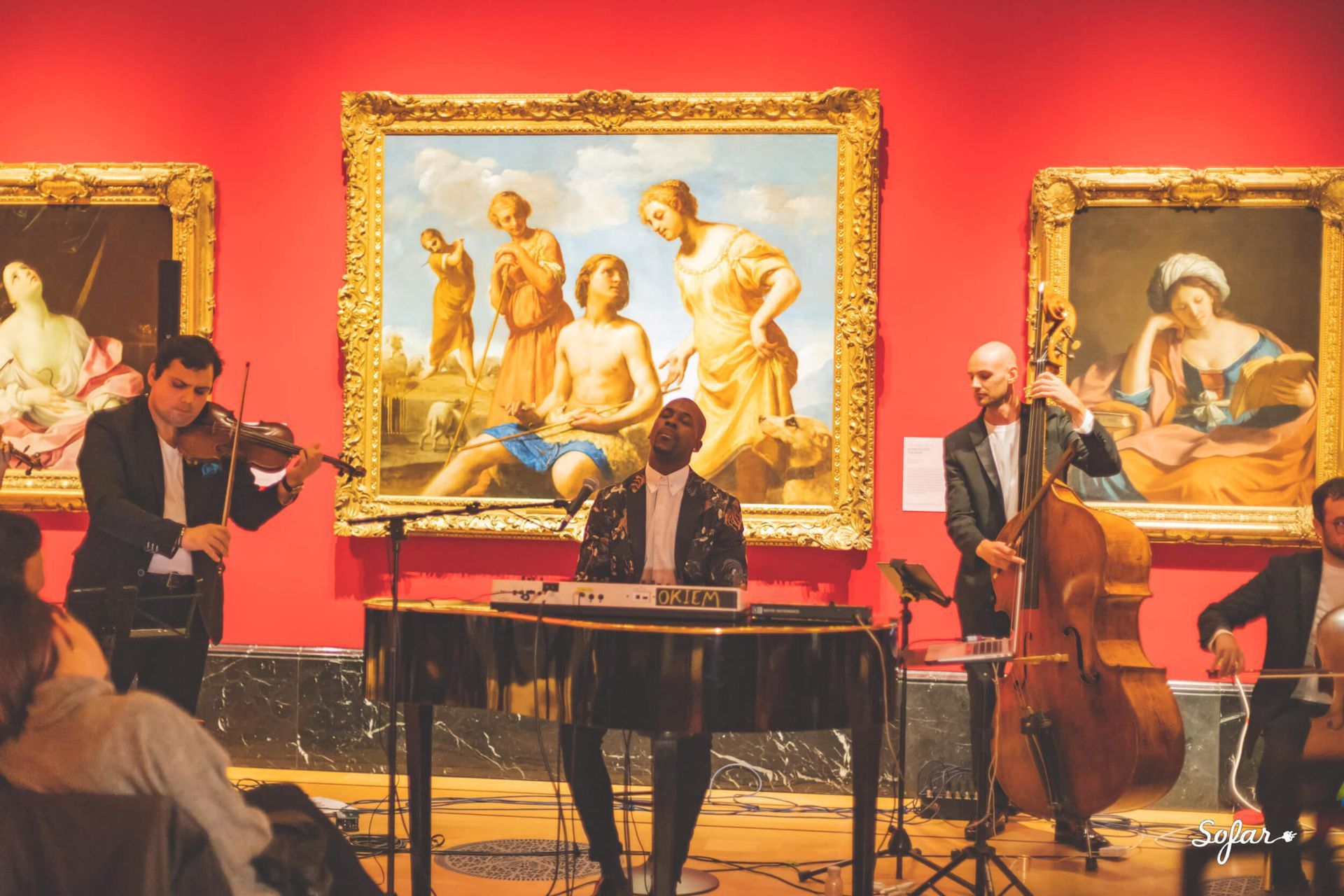 A band performs in an art museum, in front of a painting. Musicians play violin, keyboard, cello. Red walls.