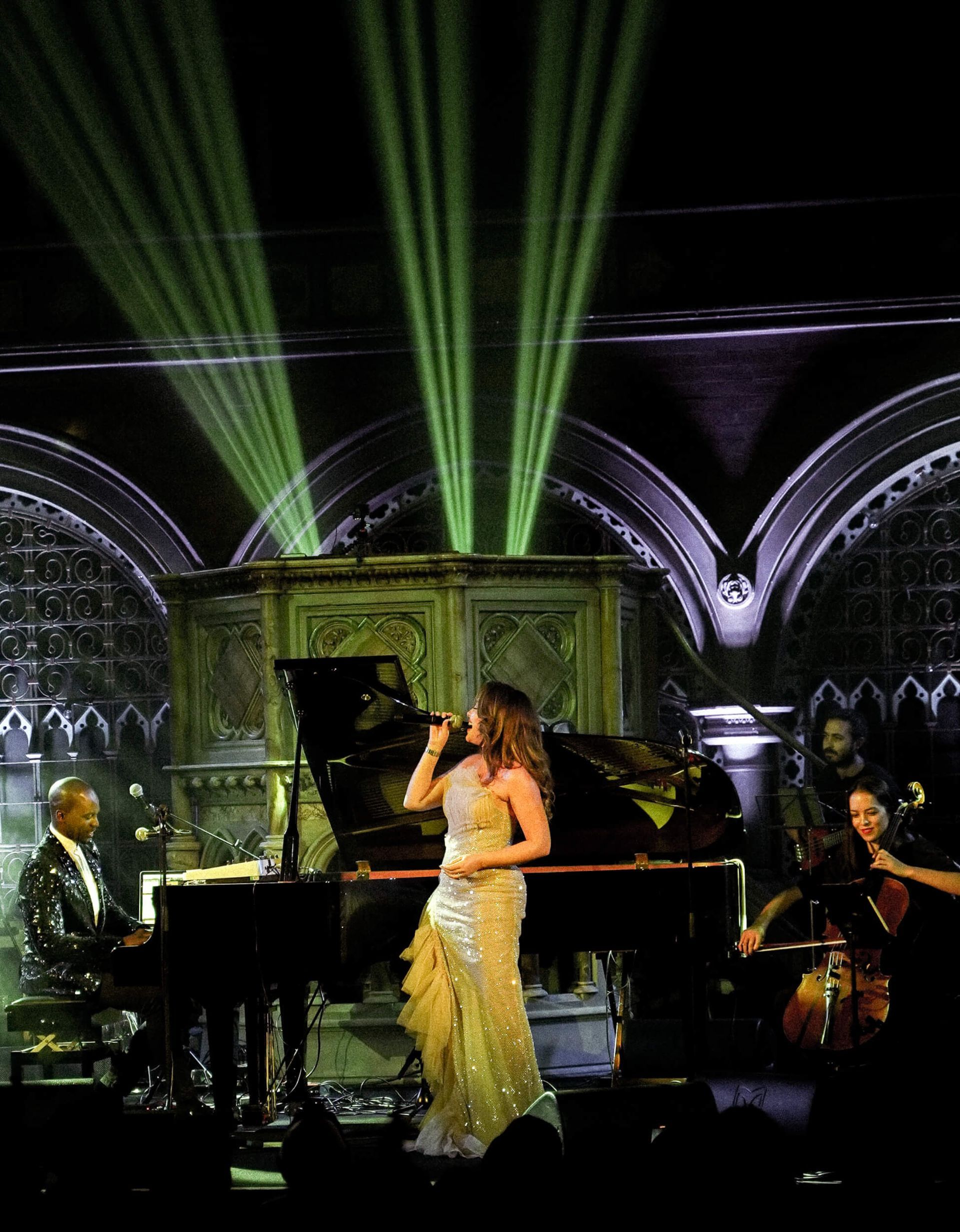 Singer in a gold gown performs with a pianist, cellist, and band on stage with spotlights.