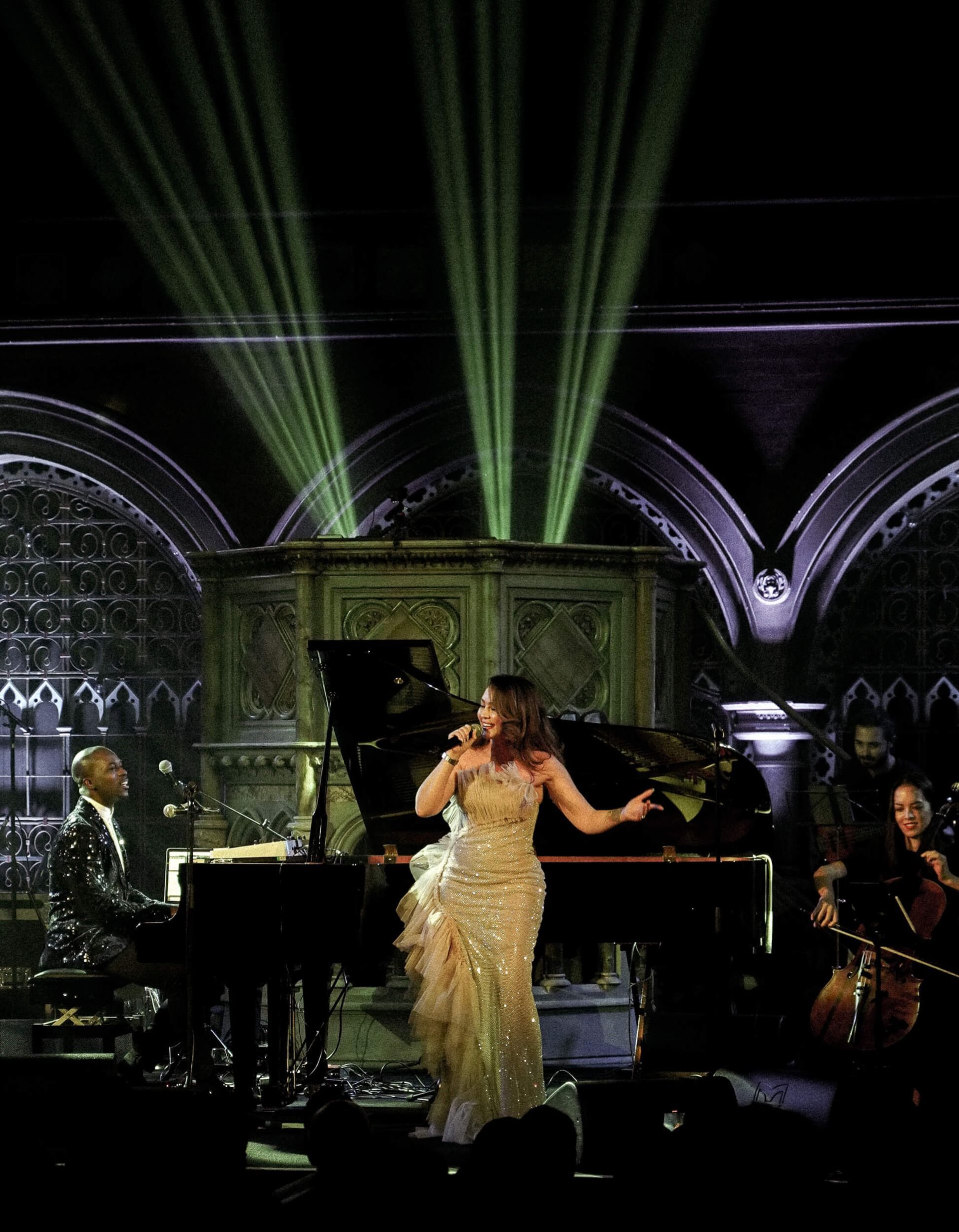 Woman in gown sings at piano with band on stage, lit by green beams.