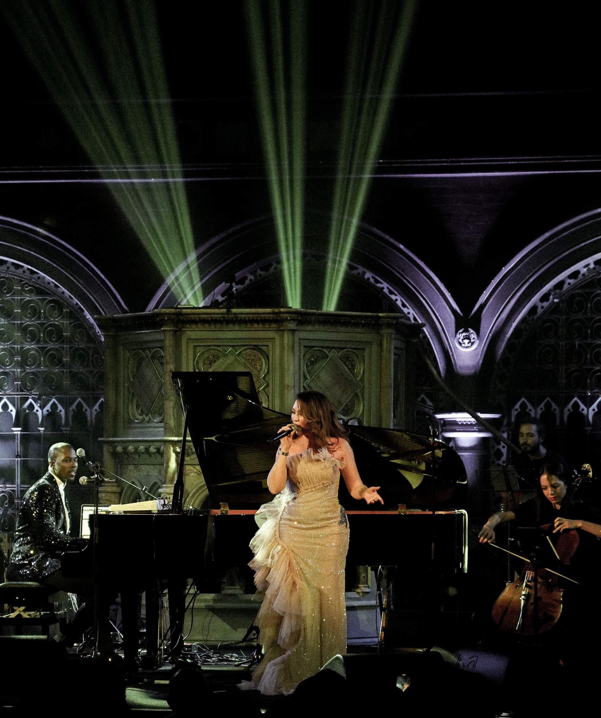 Singer in shimmering gown performs at a piano with a band on stage, green spotlights, gothic architecture.