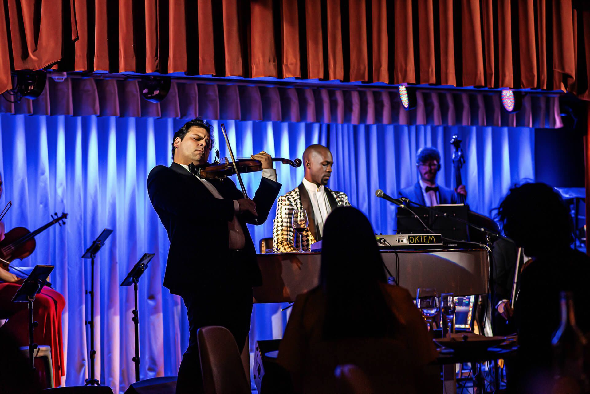 A violinist plays on stage with a band in front of an audience, stage lights.