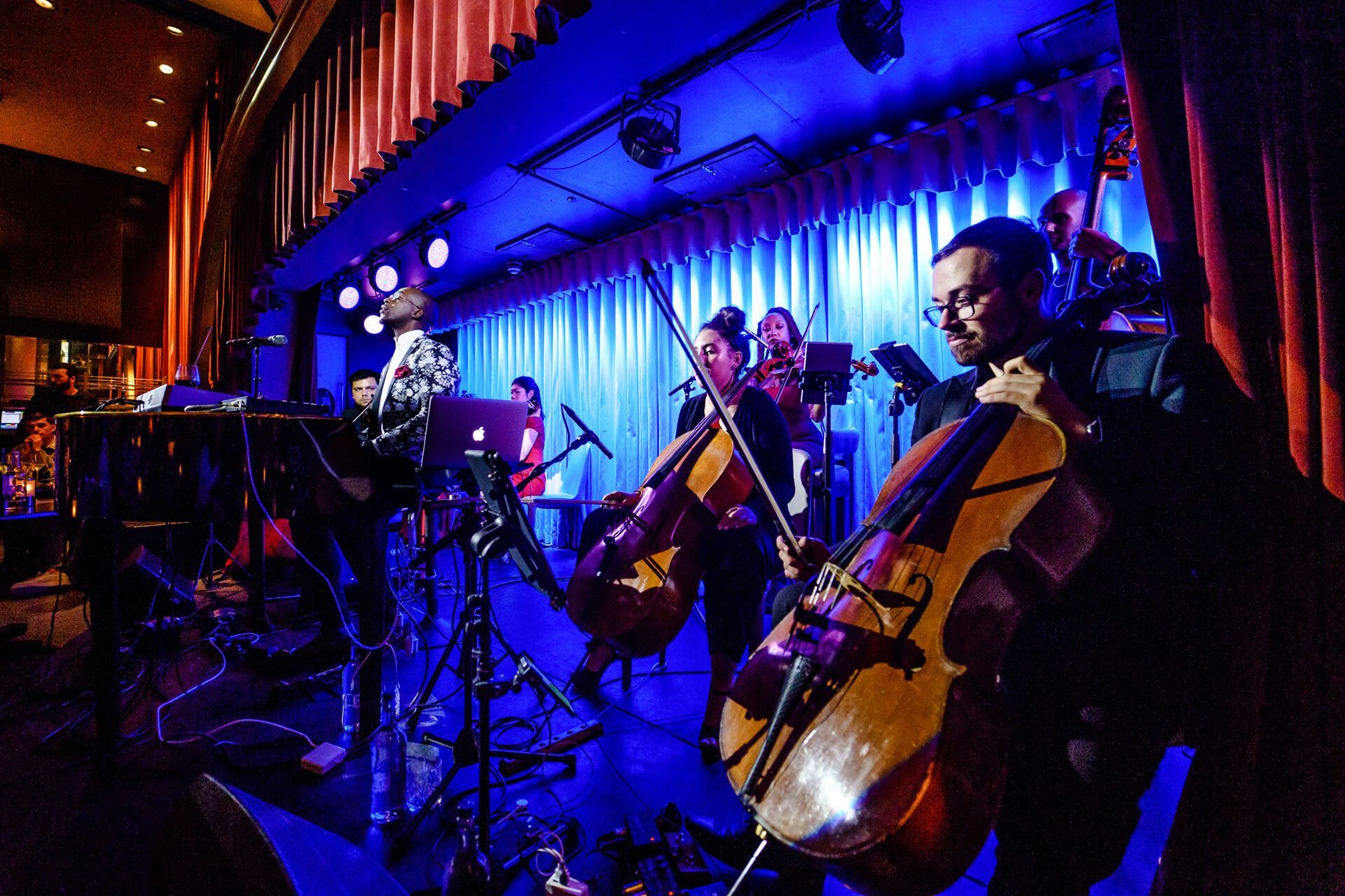 Musicians performing on a stage under blue lights. Two cellists in the foreground. Red curtains in the background.
