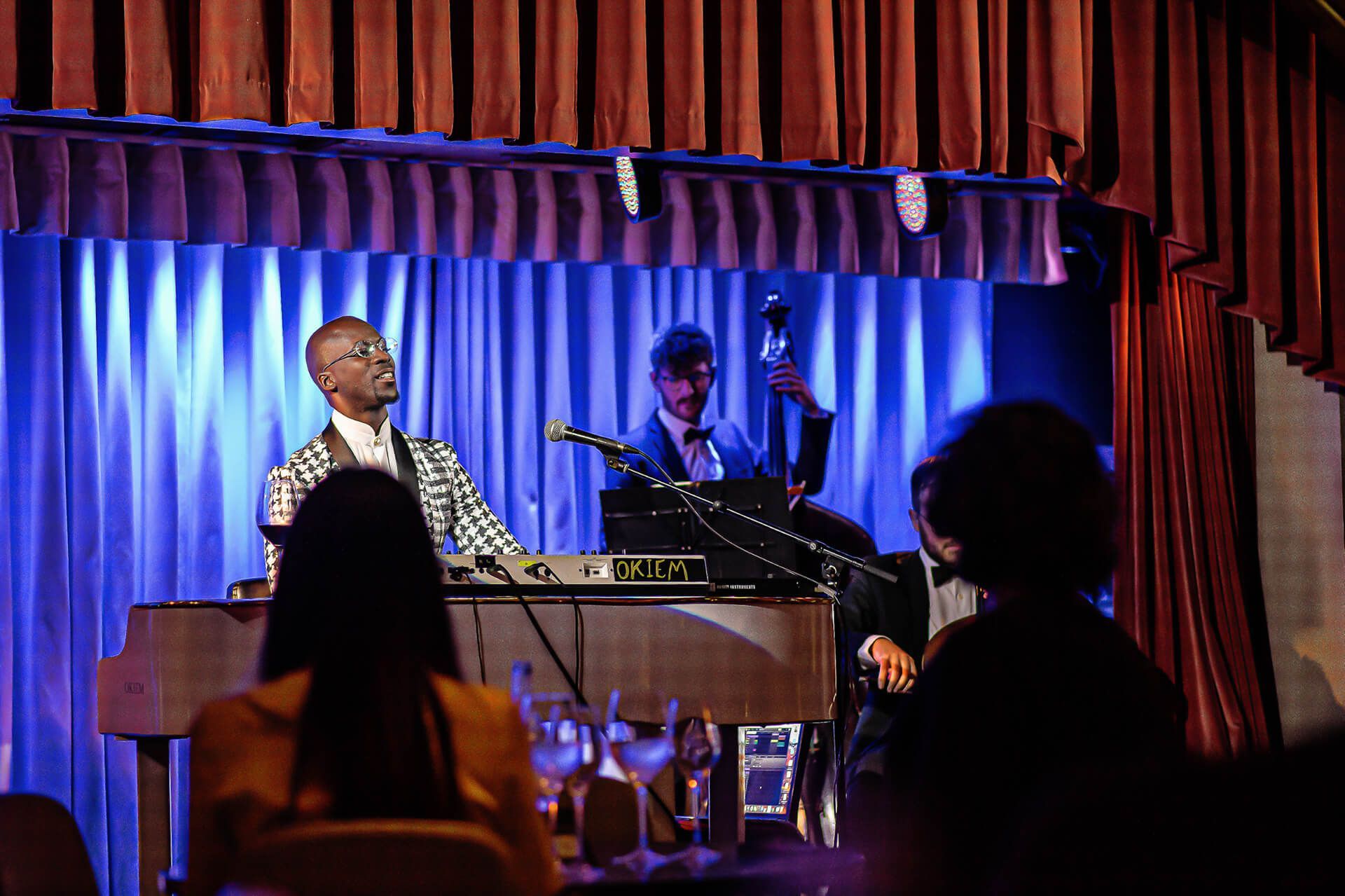 A jazz band performs on stage with a piano and upright bass. Blue and red curtains frame the musicians.