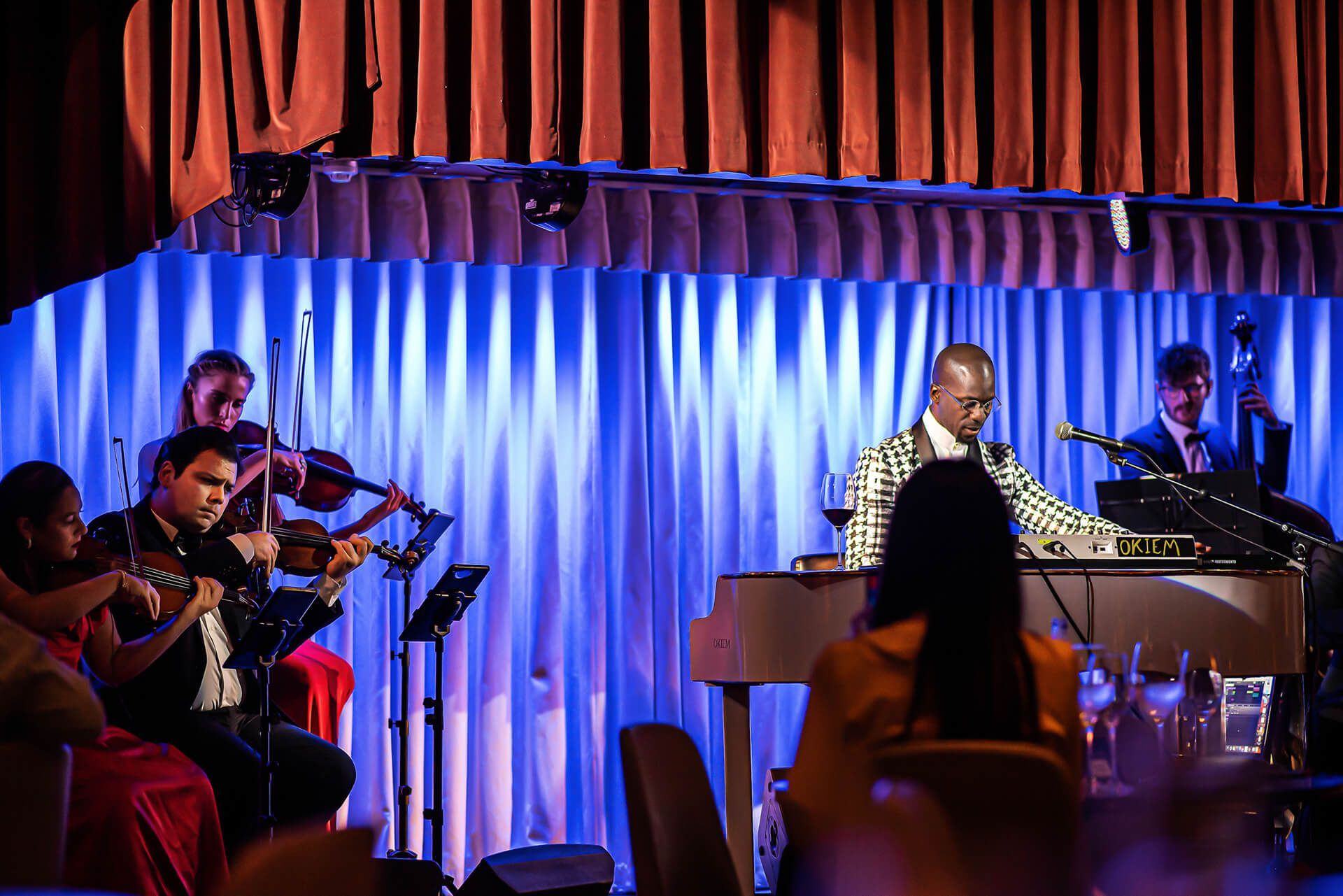 Jazz band performing on stage; piano player center, string section left, bassist right, blue backdrop, audience visible.