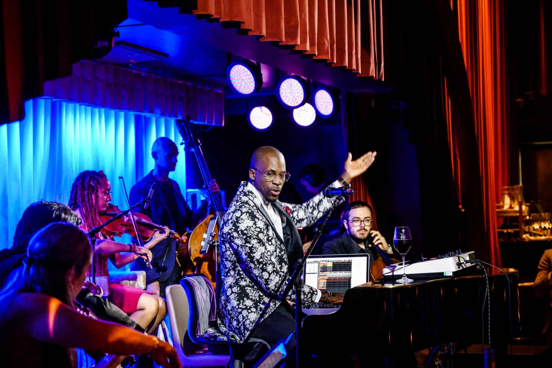 Man in patterned jacket performs on stage with band, blue lighting.