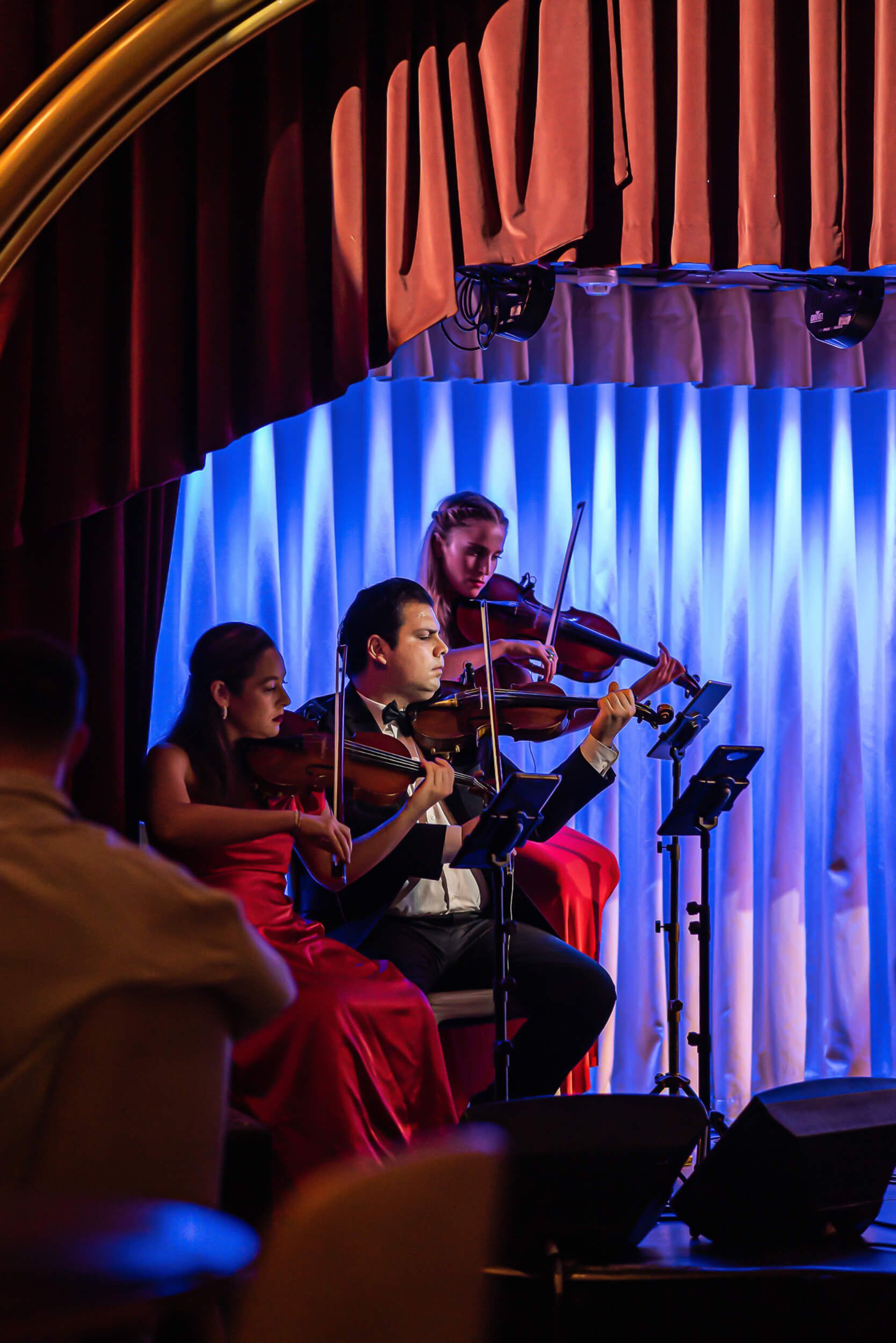Three violinists performing on a stage with blue backdrop, red curtains, and lights.