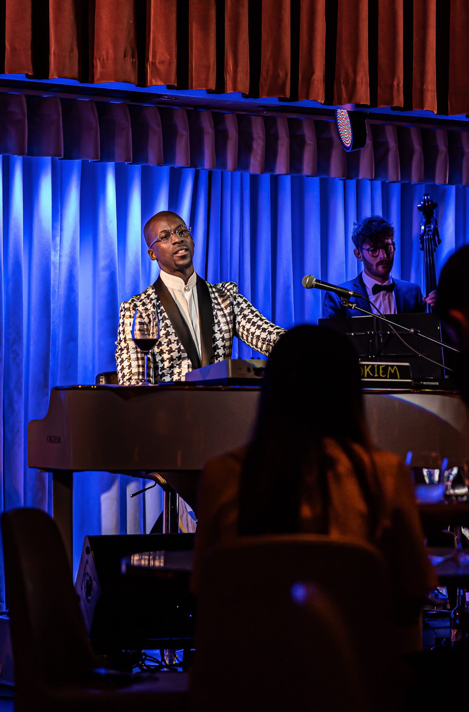Singer playing piano in a dimly lit venue, with a bassist behind.