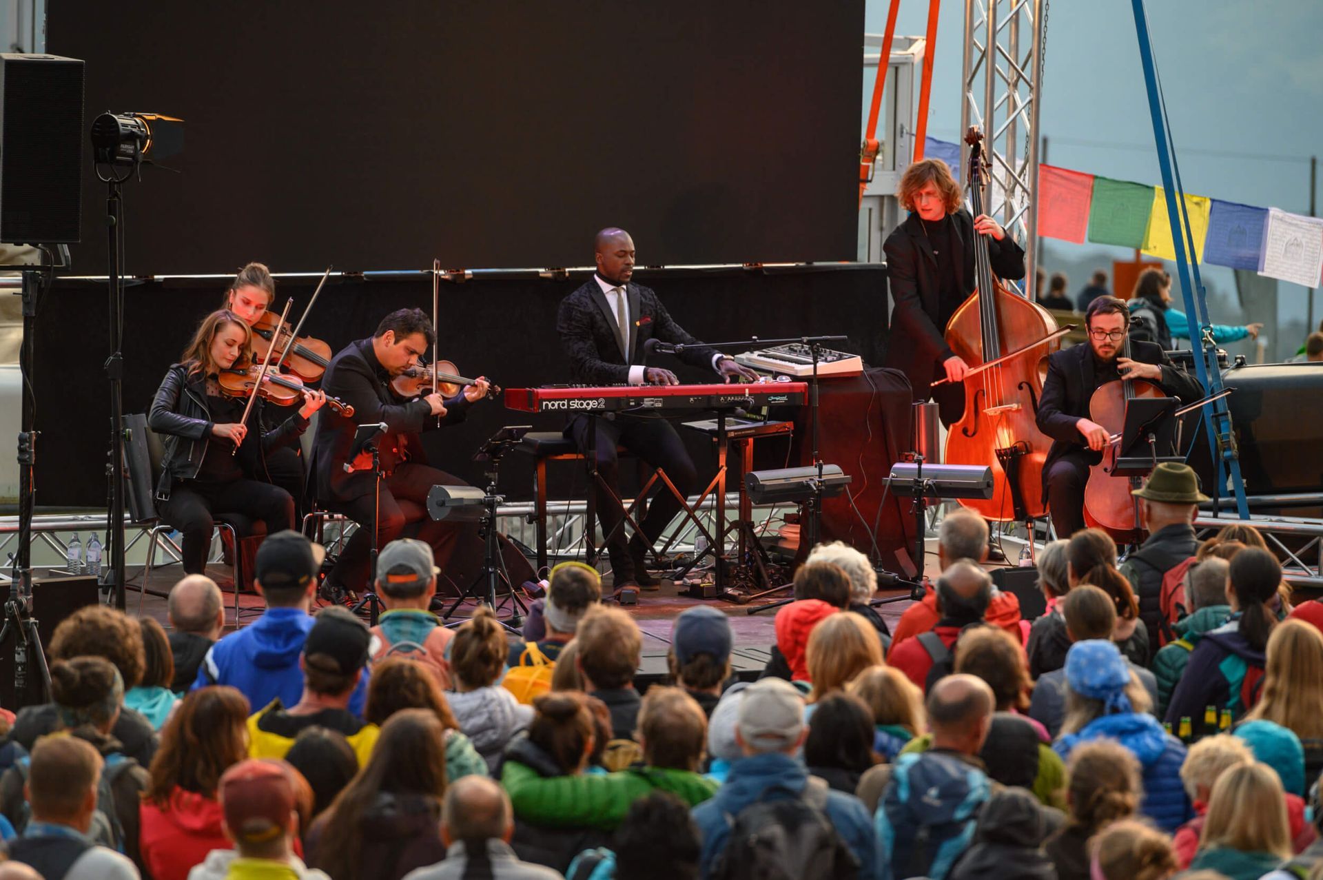 A band performs on stage for a crowd: strings, keyboard, and bass player. Outdoor setting, cloudy day.
