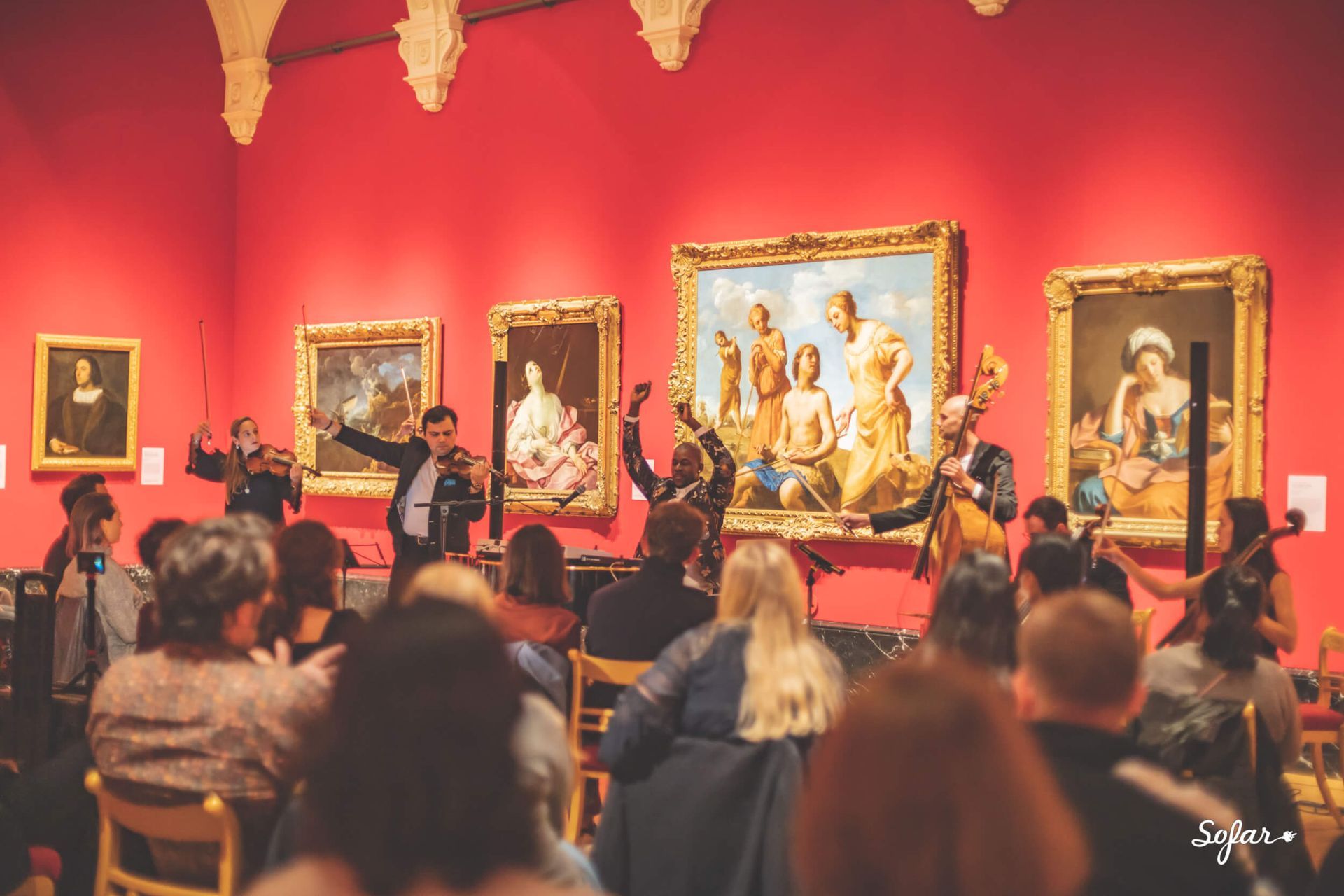 A musical ensemble performs in an art museum. Paintings hang on the red walls as the audience watches.