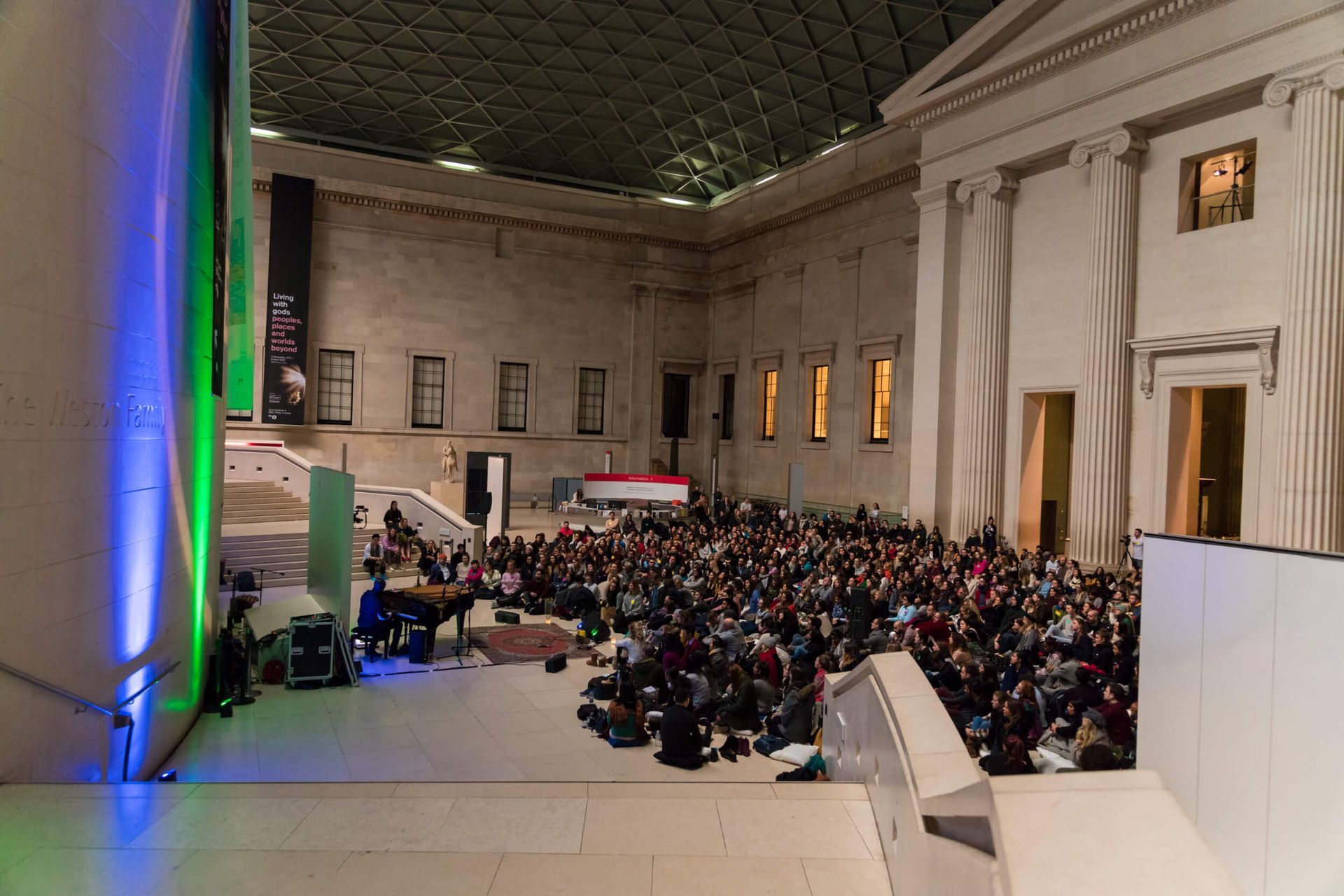 Large crowd seated in a grand hall, possibly a museum. Stage with speakers, blue and green lights, and a banner visible.