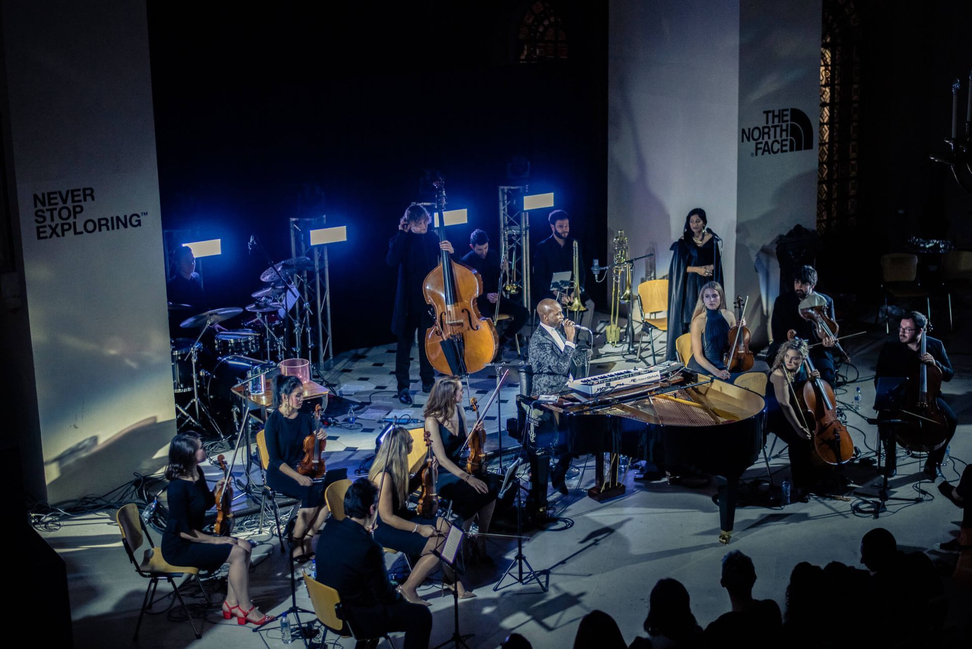 Orchestra performing on stage with a piano, various string and percussion instruments. Stage is lit with blue lights.
