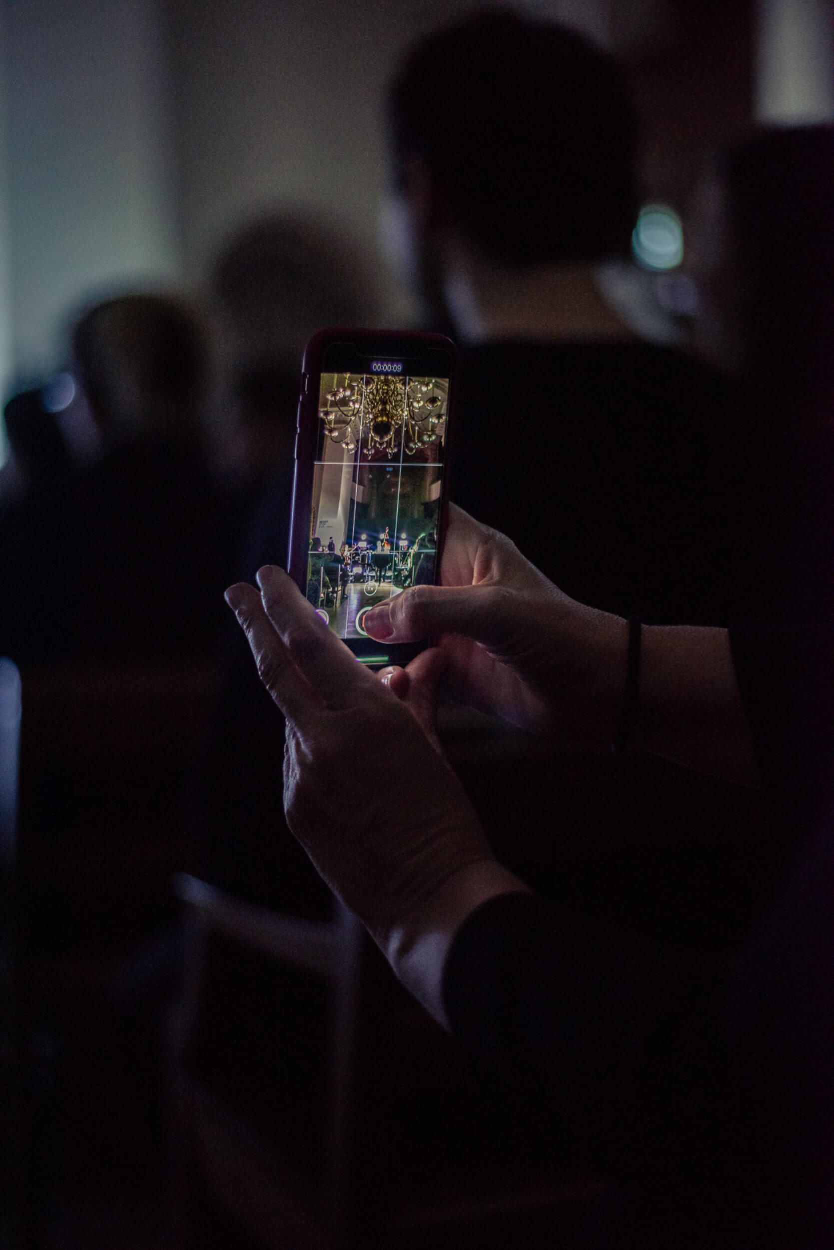 Hands holding a phone, recording an event. Bright screen illuminating the hands in a dim room.