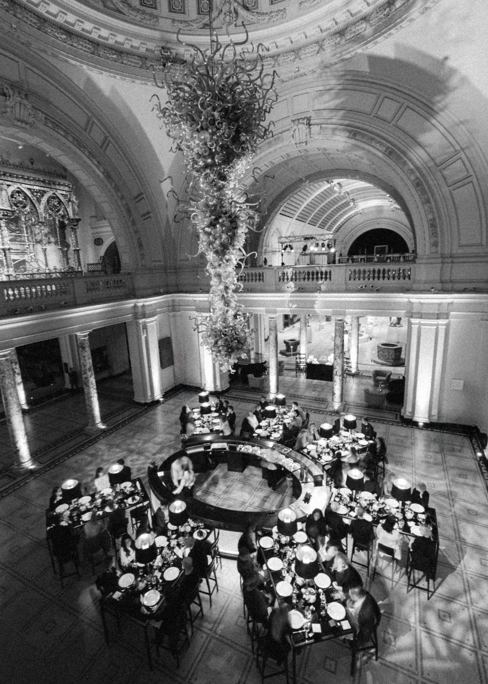 Grand hall with tables set for a meal. A tree-like sculpture hangs from the arched ceiling, with a balcony above.