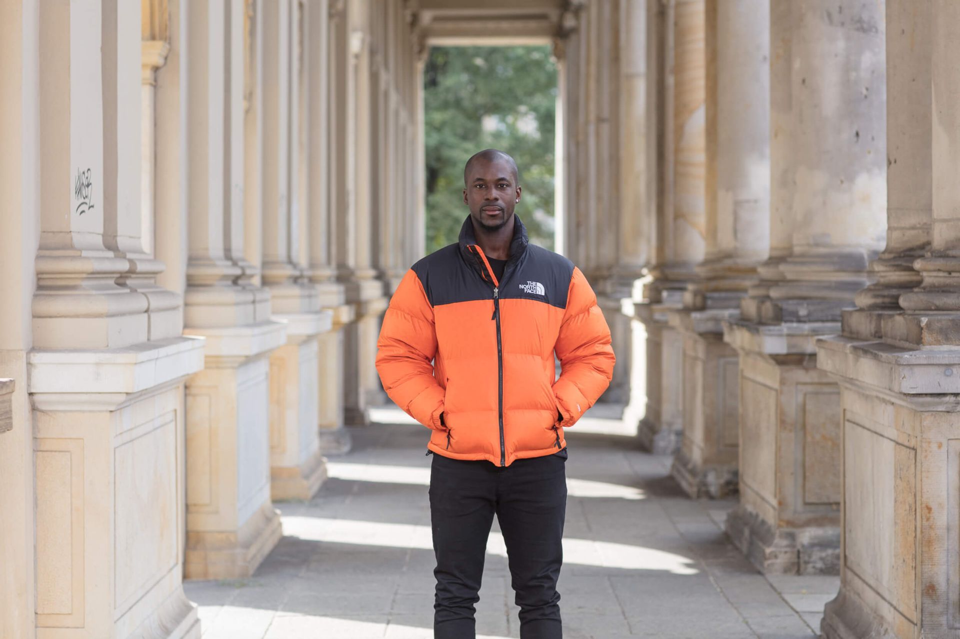 Man in orange and black puffer jacket stands in a colonnade with hands in pockets.