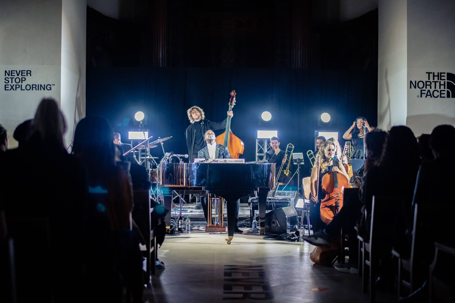 Band performing on stage with piano, upright bass, and strings. Audience in the foreground, stage is lit.