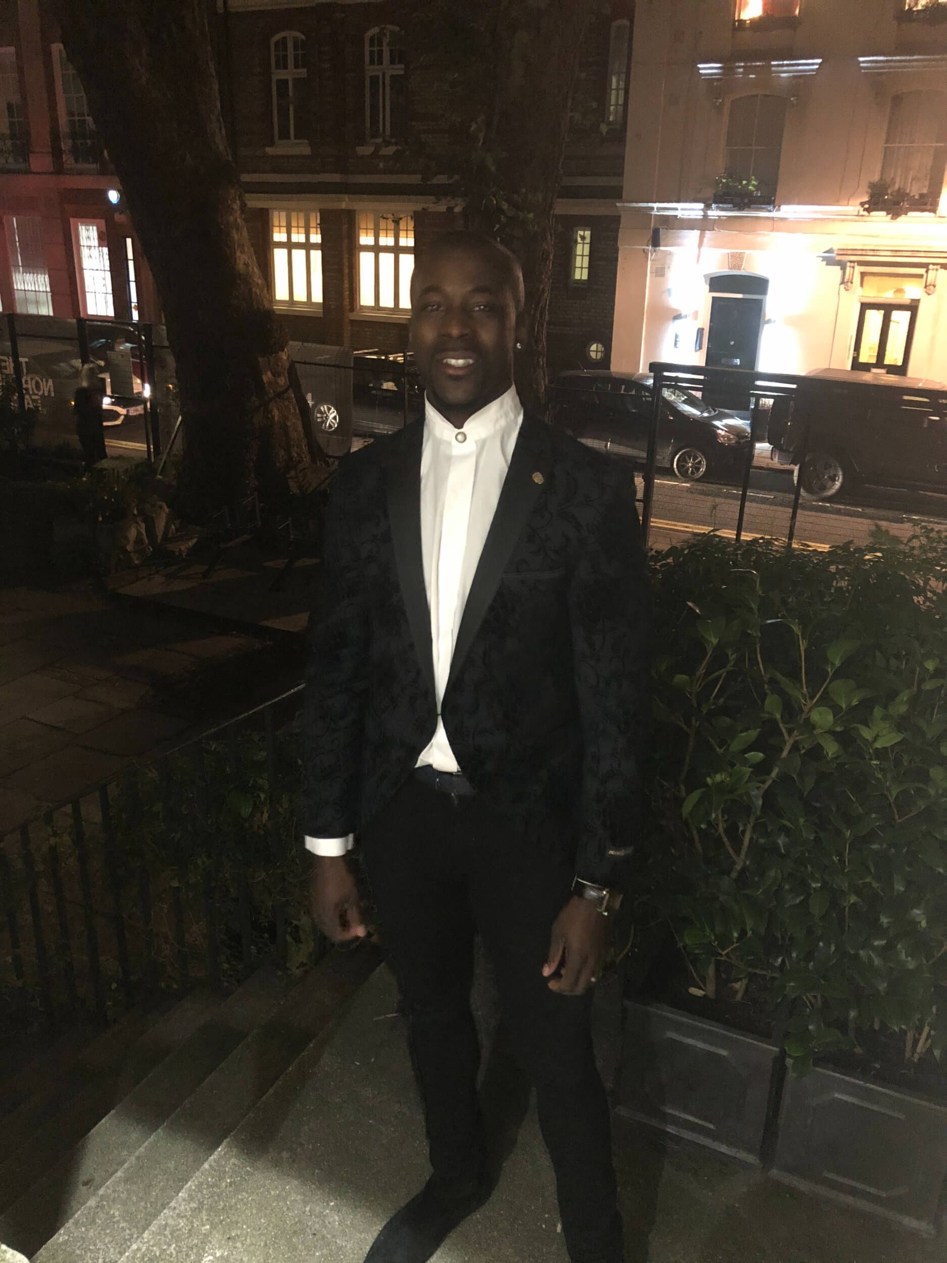 Man in a patterned black suit and white shirt, standing outside at night.