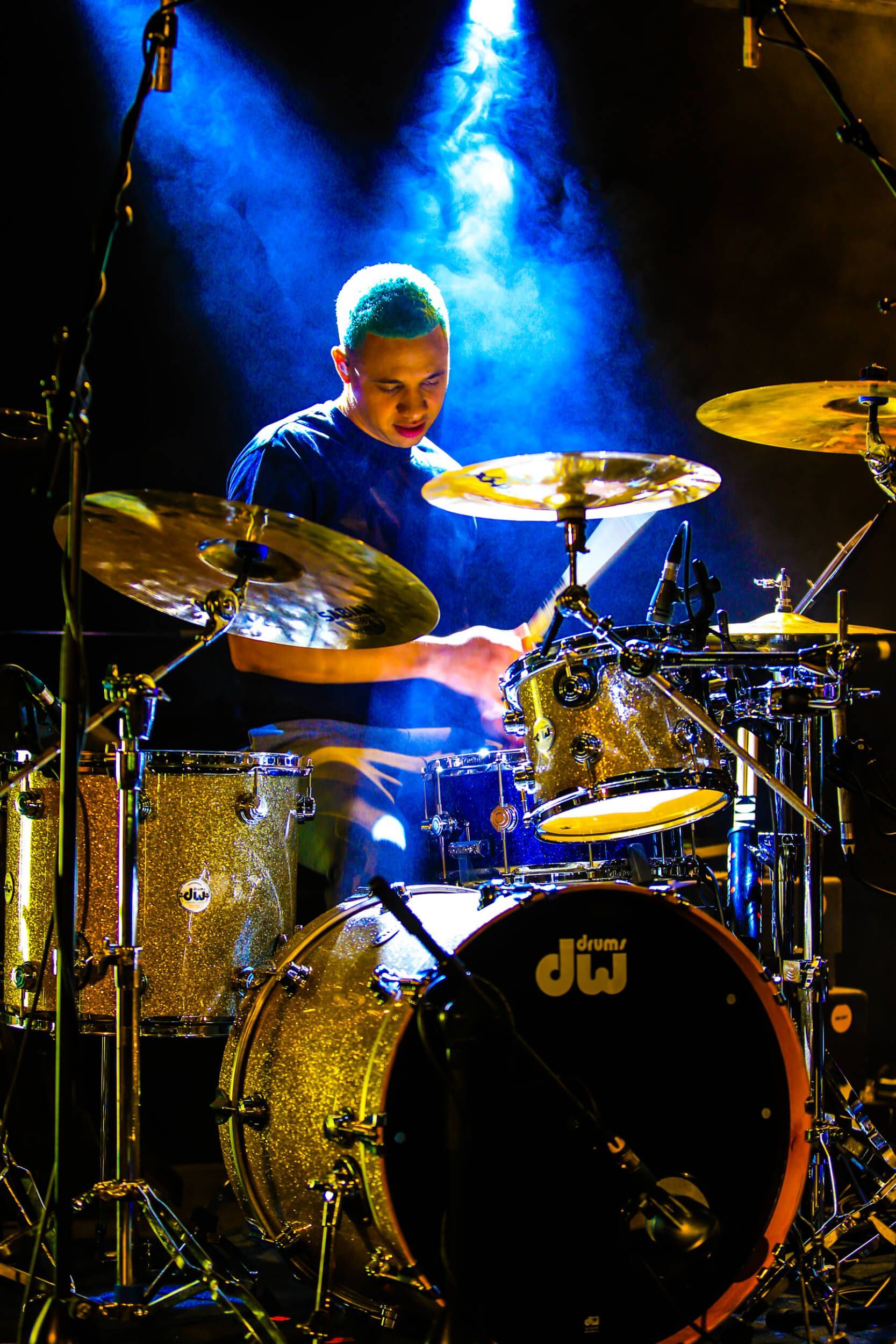Drummer playing a gold-sparkle kit under blue stage lights.