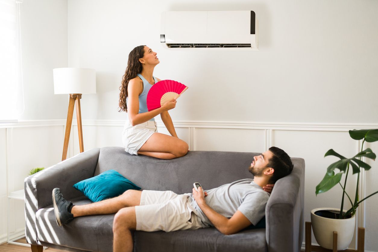 A person kneeling on a sofa uses a pink fan while looking at a wall-mounted air conditioner, as another reclines nearby.