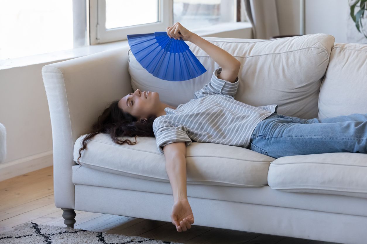 Woman lying on couch, fanning herself with a blue fan beacuse of AC cooling issues.