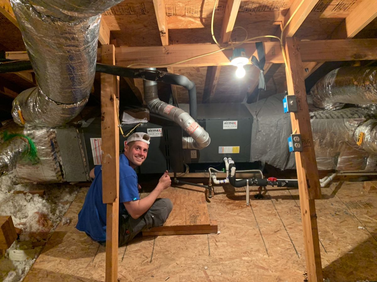 A smiling man inspects HVAC system in an attic, with ductwork and a lightbulb visible.