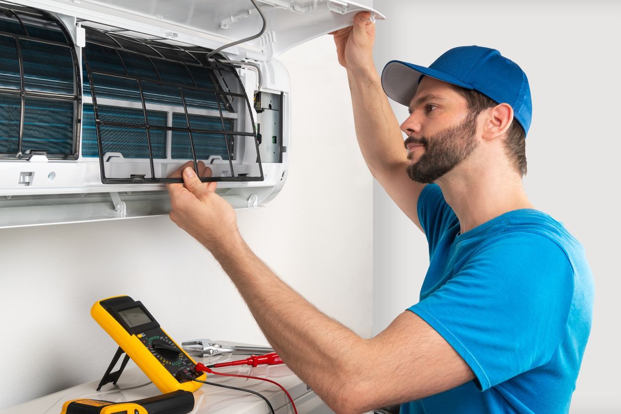 Man in blue uniform inspecting an air conditioner with a multimeter in a white room.