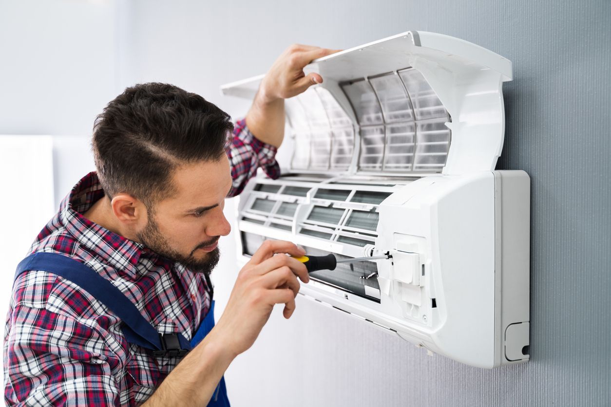 A technician in a plaid shirt and blue overalls uses a screwdriver to repair a wall-mounted air conditioning unit.