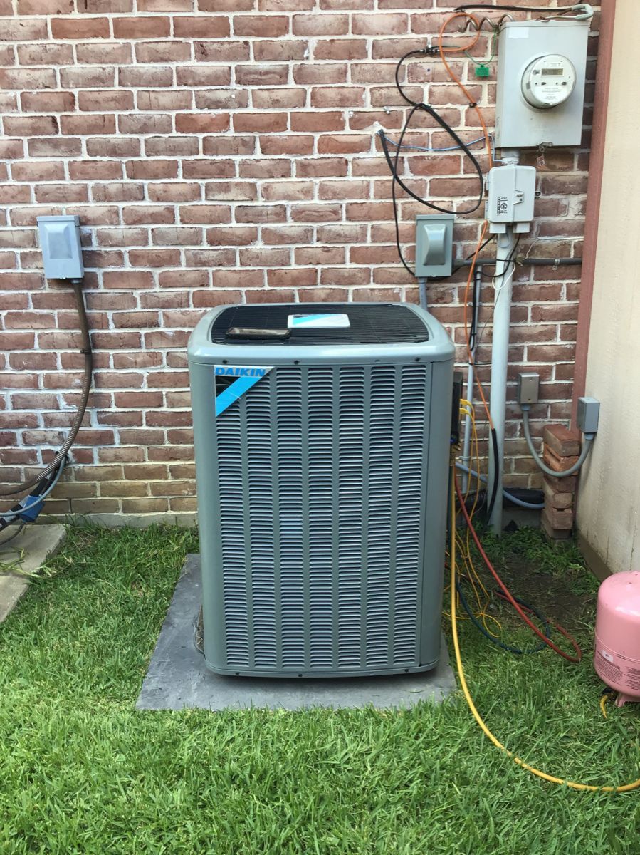Air conditioning unit against a brick wall, in a grassy area with electrical components.
