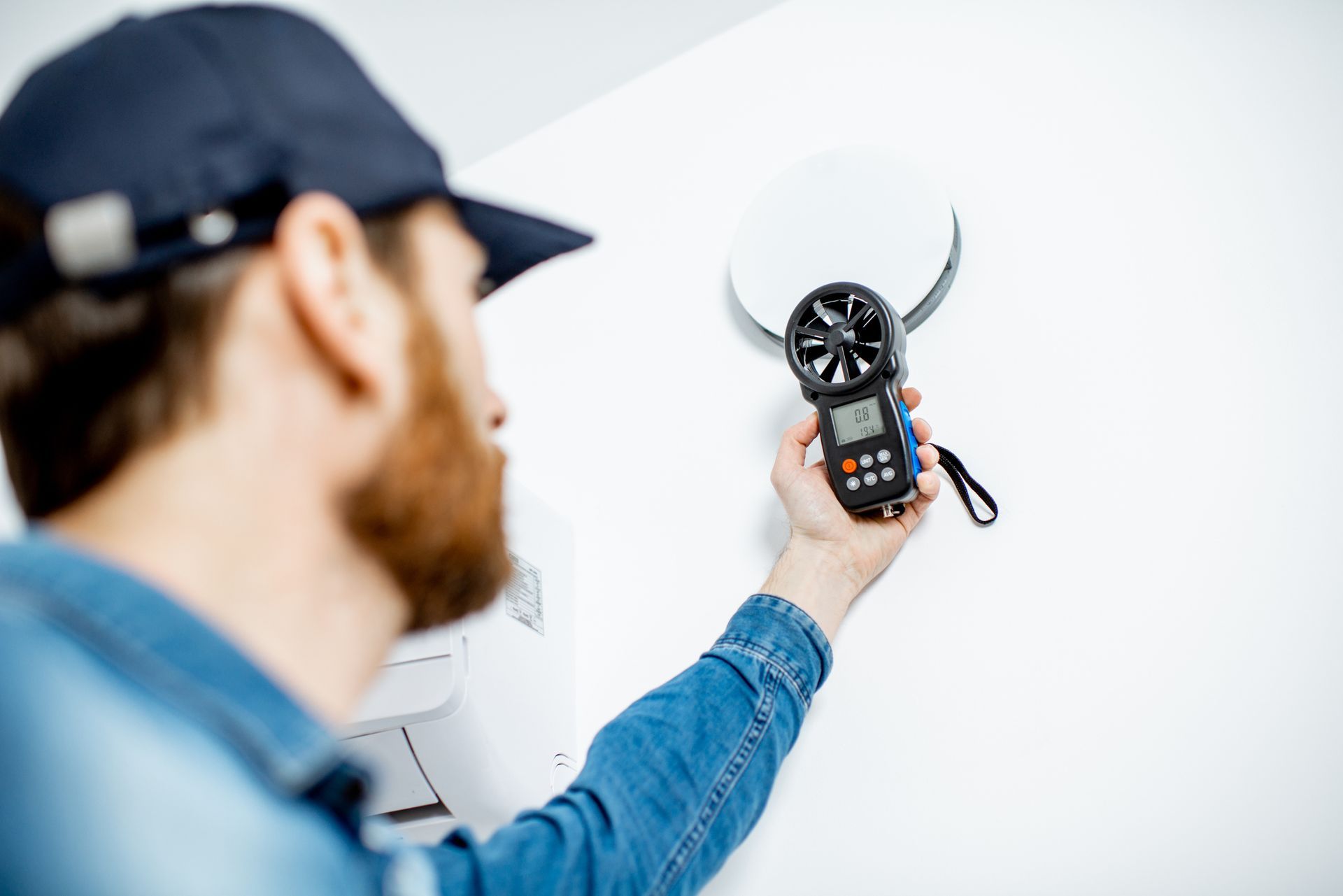 Man in blue shirt and cap uses anemometer on a white vent cover.