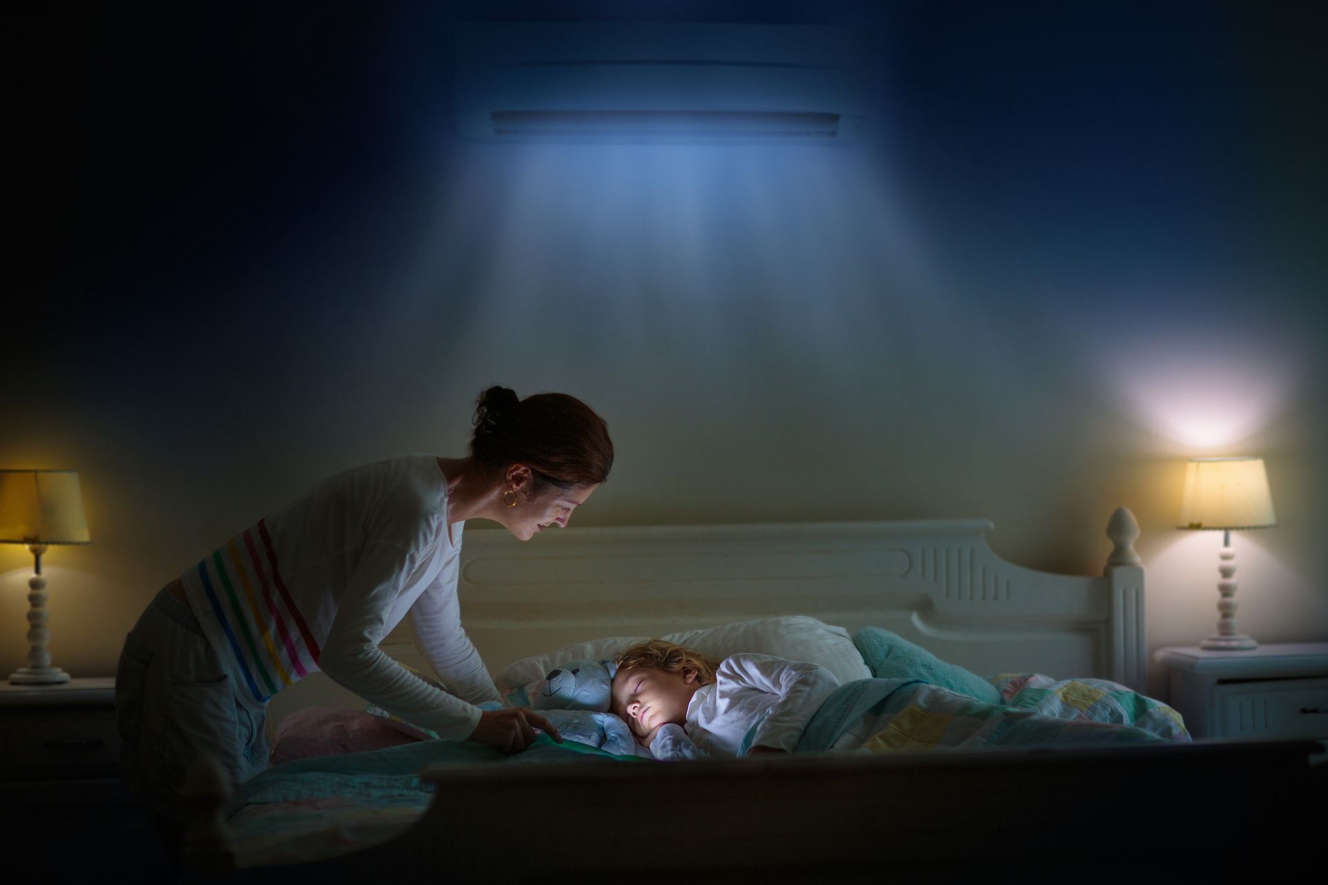 Woman tucking a child into bed in a dim bedroom, lit by bedside lamps and soft overhead light