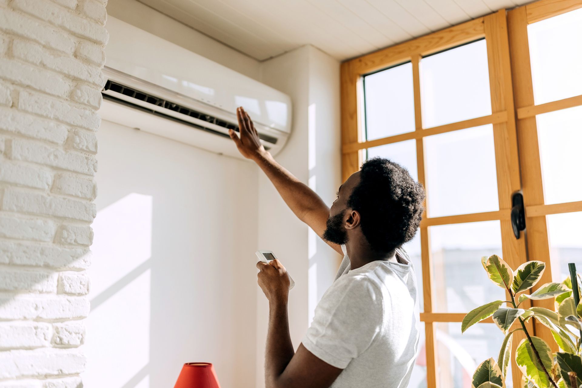 A person reaches towards a wall-mounted air conditioner unit indoors. Bright light comes in the window.