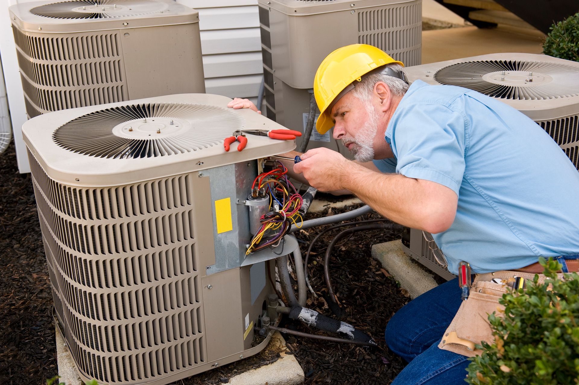 Technician in yellow hard hat repairing outdoor HVAC unit with exposed wiring.
