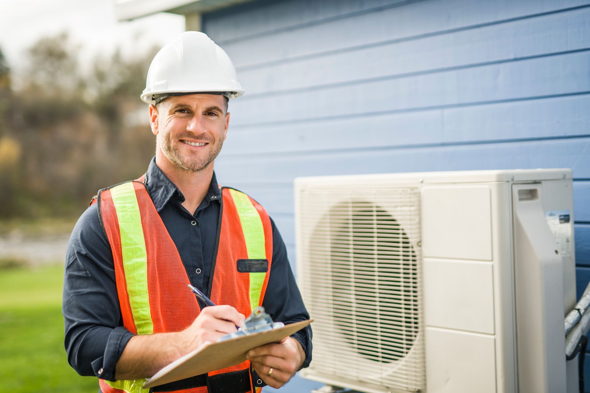 Technician in hard hat and safety vest inspecting an outdoor HVAC unit with a clipboard.