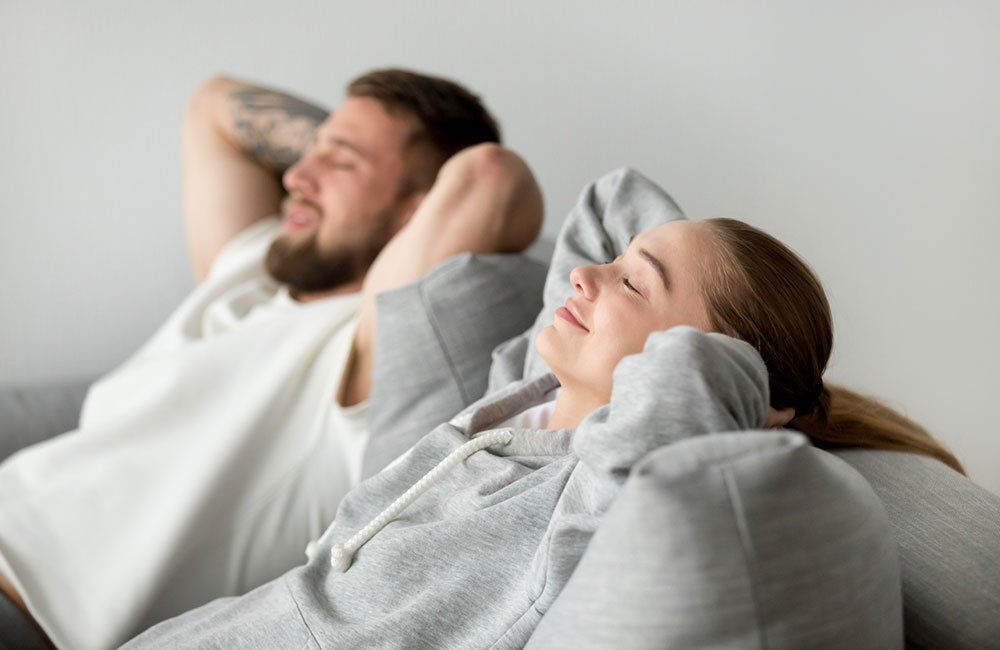 Couple relaxing on a couch, eyes closed, arms behind their heads, smiling, light gray setting.