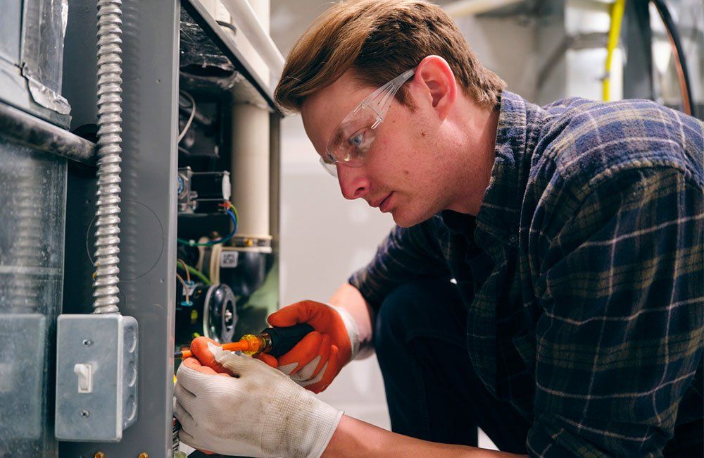 Airmax Air Conditioning & Heating technician in plaid shirt and safety glasses, inspecting furnace wiring.