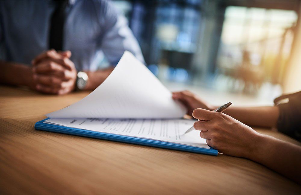 Person signing a document on a clipboard, sitting across from another person at a table indoors.
