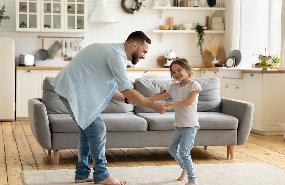Father and daughter dancing together in their living room, both smiling.