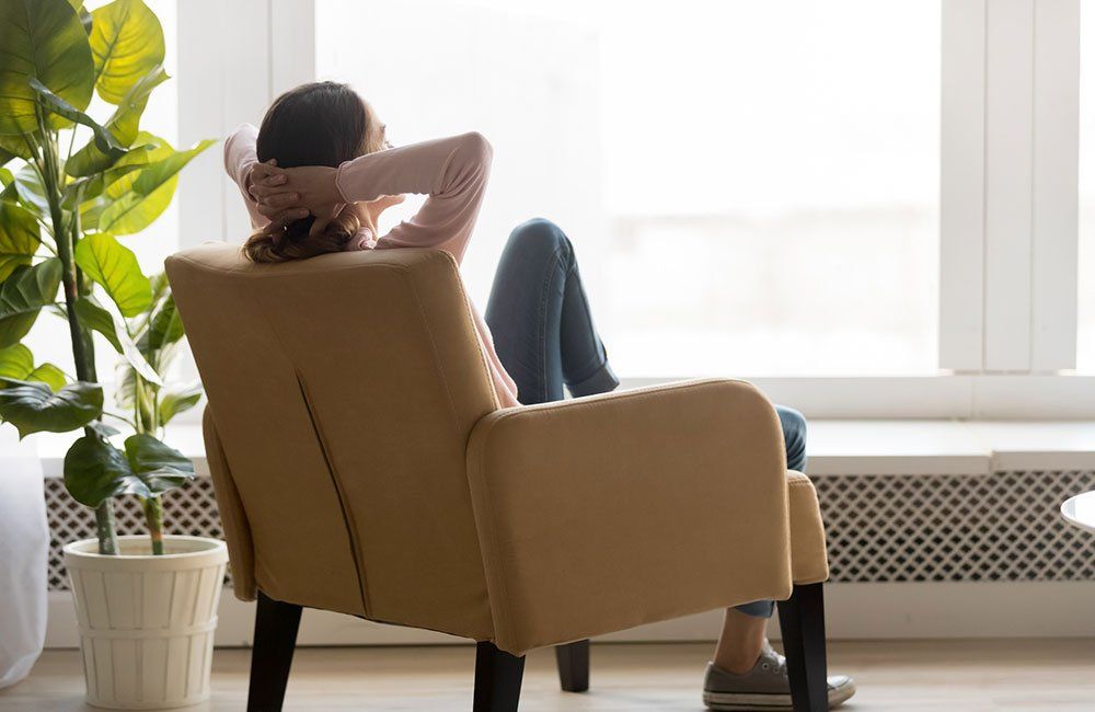 Woman relaxing in a yellow armchair, looking out a window in a bright room.