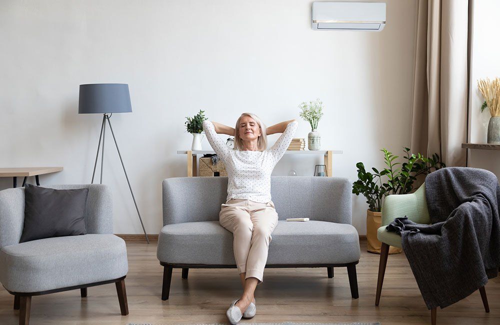 Woman relaxing on a gray sofa under AC, hands behind head, in a bright living room.