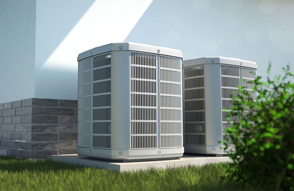Two air conditioning units outside a building on a concrete pad, surrounded by grass and a bush.