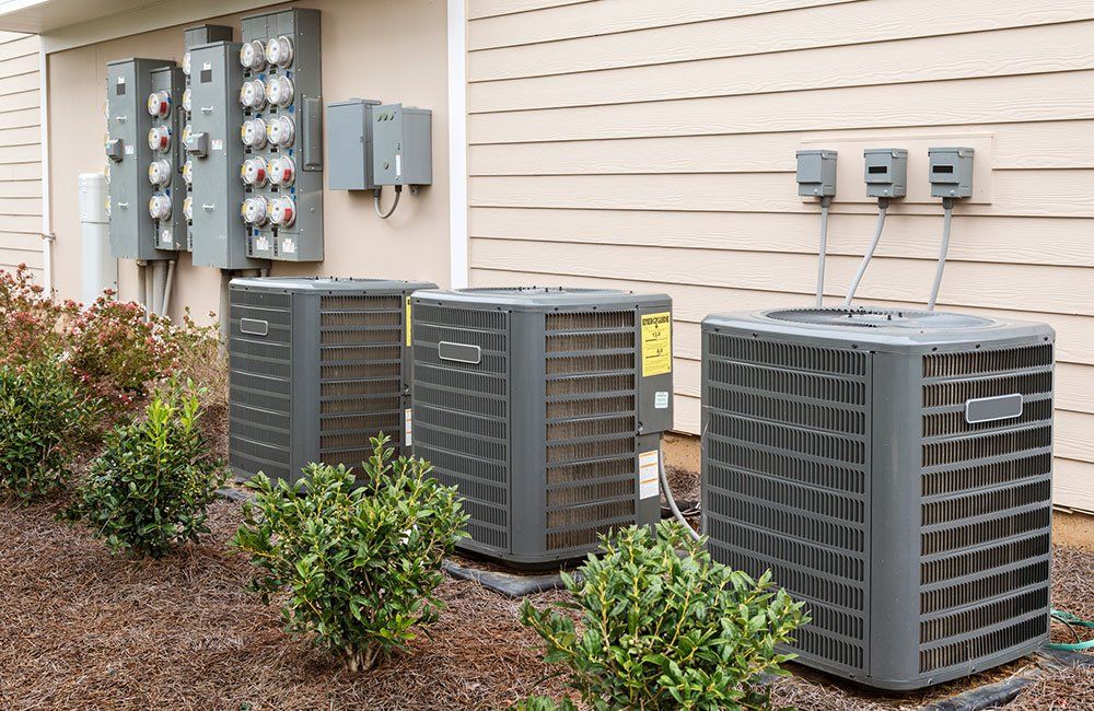 Three air conditioning units next to a building with electrical meters and bushes.